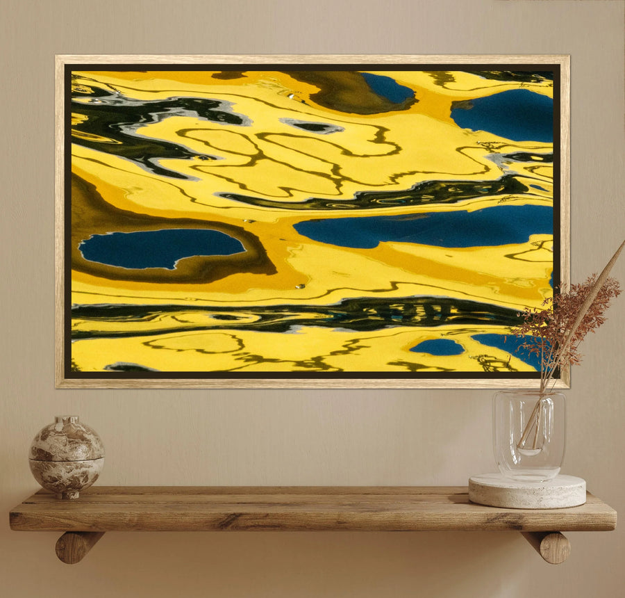 Abstract framed wall art featuring bold yellow, black, and deep blue marbled patterns, displayed above a wooden shelf with decorative objects in a styled interior setting.