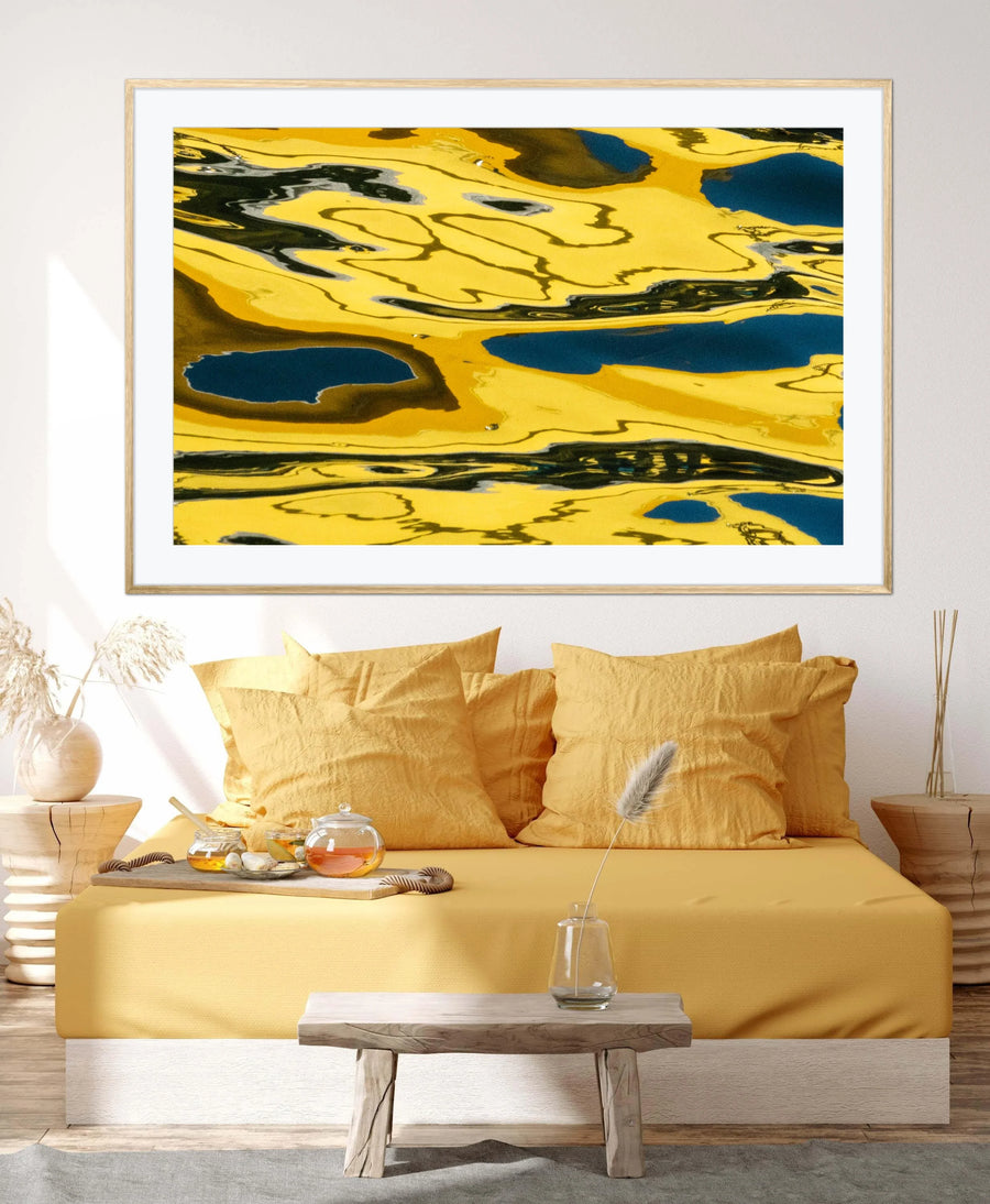 A large framed abstract art print featuring a yellow, black, and dark blue marbled pattern, displayed on a white wall above a modern yellow bed in a styled bedroom setting.