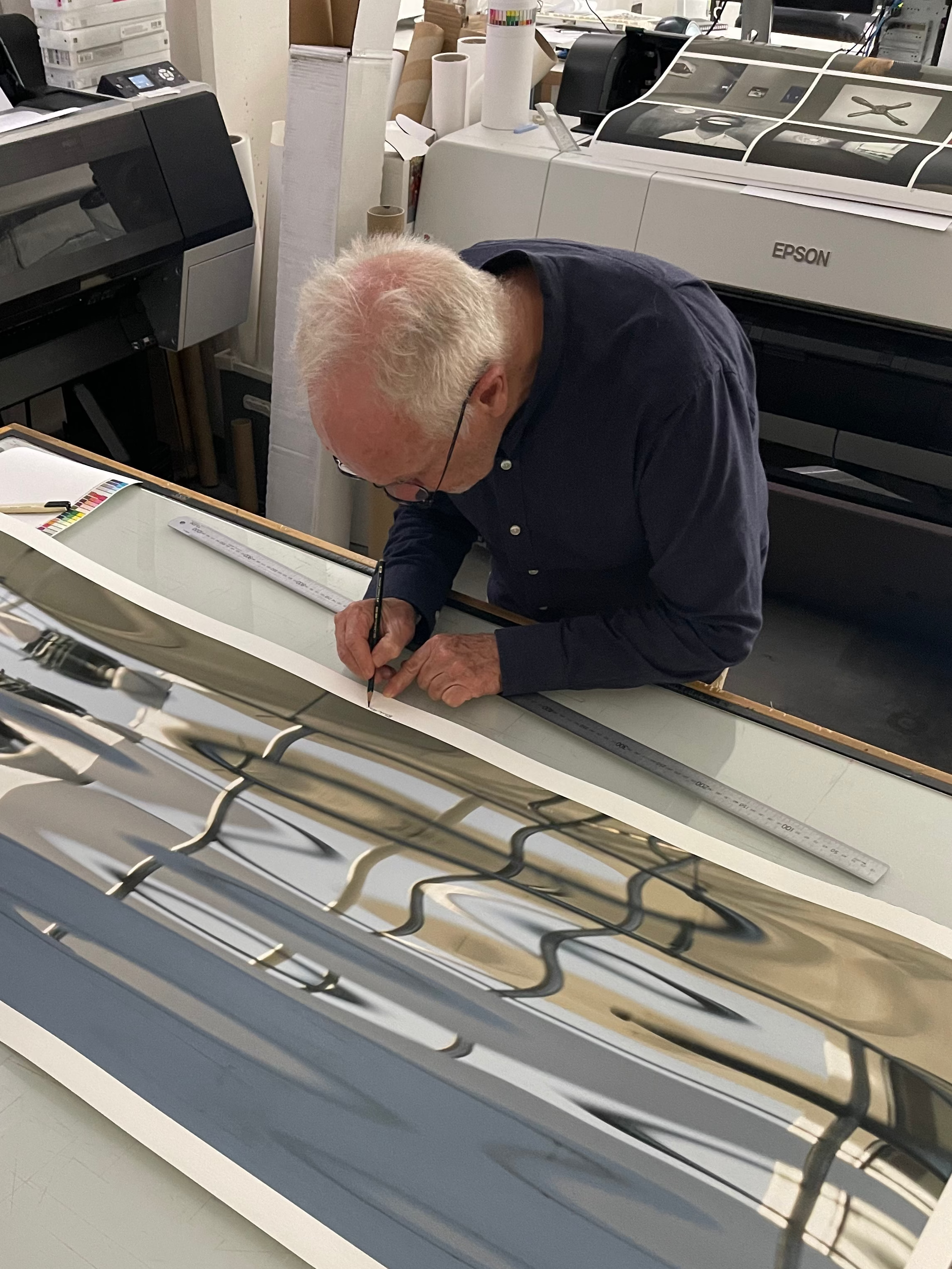 An artist in a dark navy shirt is carefully tracing or drawing on a large, wide-format glossy art print laid out on a table in a studio. The print features an abstract design with a prominent slate blue-gray section and a reflective silver-gray section.