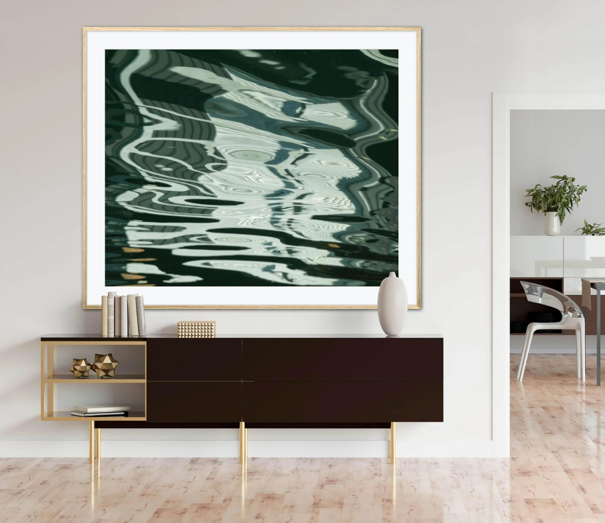 A large framed abstract art print featuring dark green and white water-like reflections hangs on a white wall above a dark brown sideboard with gold legs in a modern living room setting.