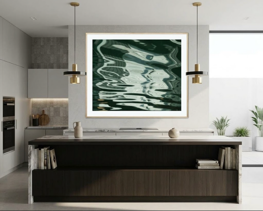 A large framed abstract art print featuring dark green and teal water-like ripples, displayed on a white wall above a modern kitchen island in a bright, contemporary kitchen setting.
