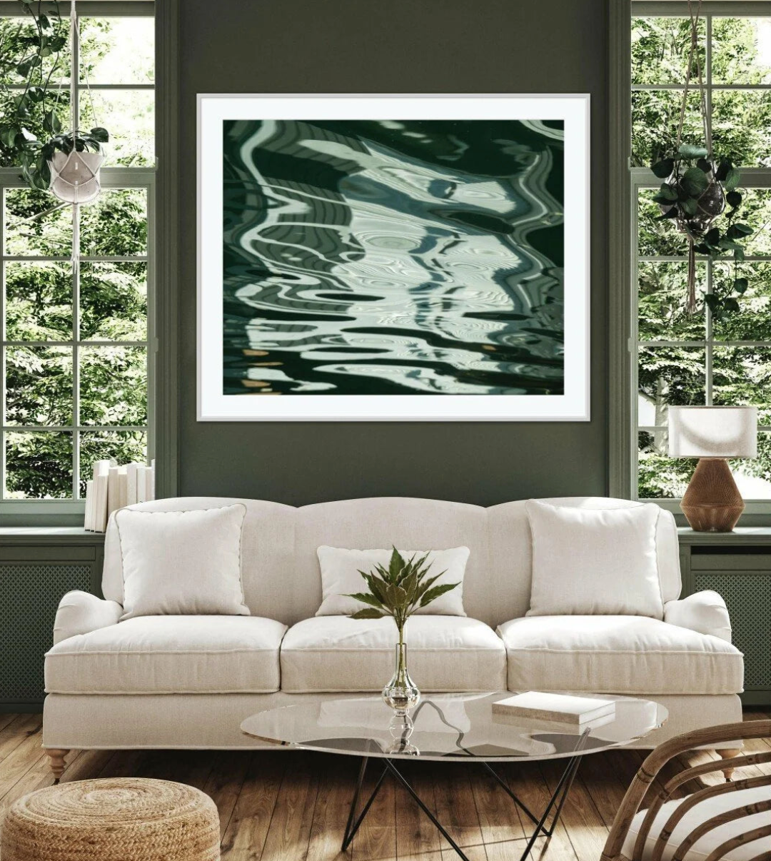 A framed abstract art print featuring dark green and teal water-like reflections, displayed on a dark green wall above a cream-colored sofa in a modern living room setting.