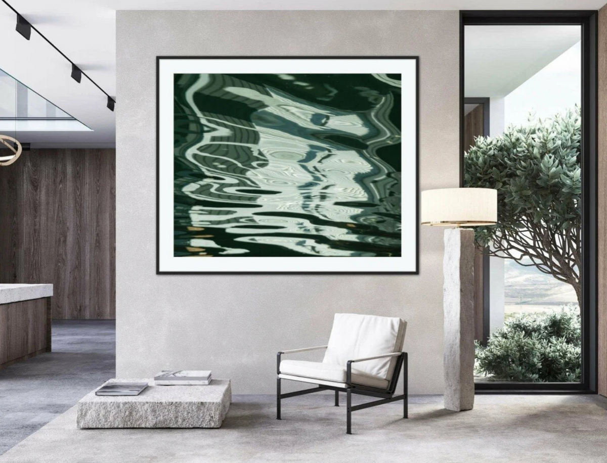 A framed abstract art print featuring dark green and black wavy reflections, displayed on a light gray wall in a modern living room setting with a white armchair, stone coffee table, and floor lamp.
